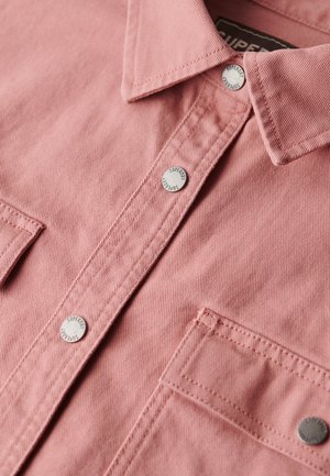 Chemise rose clair en tissu texturé, avec un col pointu et des boutons-pression en métal argenté ornés du logo "Superdry" en relief. Deux poches poitrine.