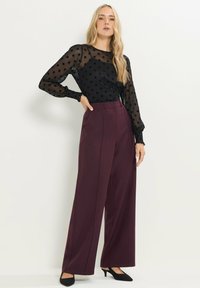 Zwarte doorschijnende blouse met stippen, gecombineerd met hoge taille, wijde bordeauxrode broeken. Zwarte puntige hakken maken de outfit af.