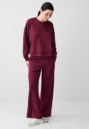 Kobieta w luźnej bordowej bluzie i spodniach o szerokich nogawkach, patrząca w dół, z rękami w kieszeniach, białe trampki na jednolitym tle.