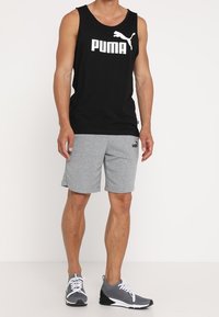 Musta hihaton toppi, jossa valkoinen Puma-logo, yhdistettynä harmaisiin shortseihin. Asu viimeistelevät harmaat urheilujalkineet, joissa on verkkokoristeet.