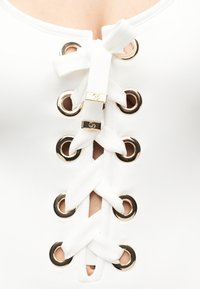 Λευκό ένδυμα με χρυσά μεταλλικά ραφές και φαρδιές κορδέλες δεμένες σε φιόγκο, αποκαλύπτοντας μικρές εμφανίσεις δέρματος κάτω από το δέσιμο στο μπροστινό μέρος.