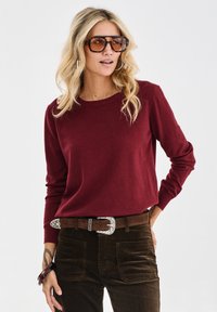 Burgunderfarbener Pullover mit langen Ärmeln und runder Ausschnitt, kombiniert mit braunen Cordhosen und einem Gürtel mit einer silberfarbenen Schnalle.