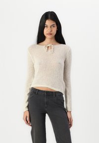 Lys beige strikket sweater med lange ærmer og rund halsudskæring, der har en sløjfedetalje foran; parret med mørkegrå jeans.