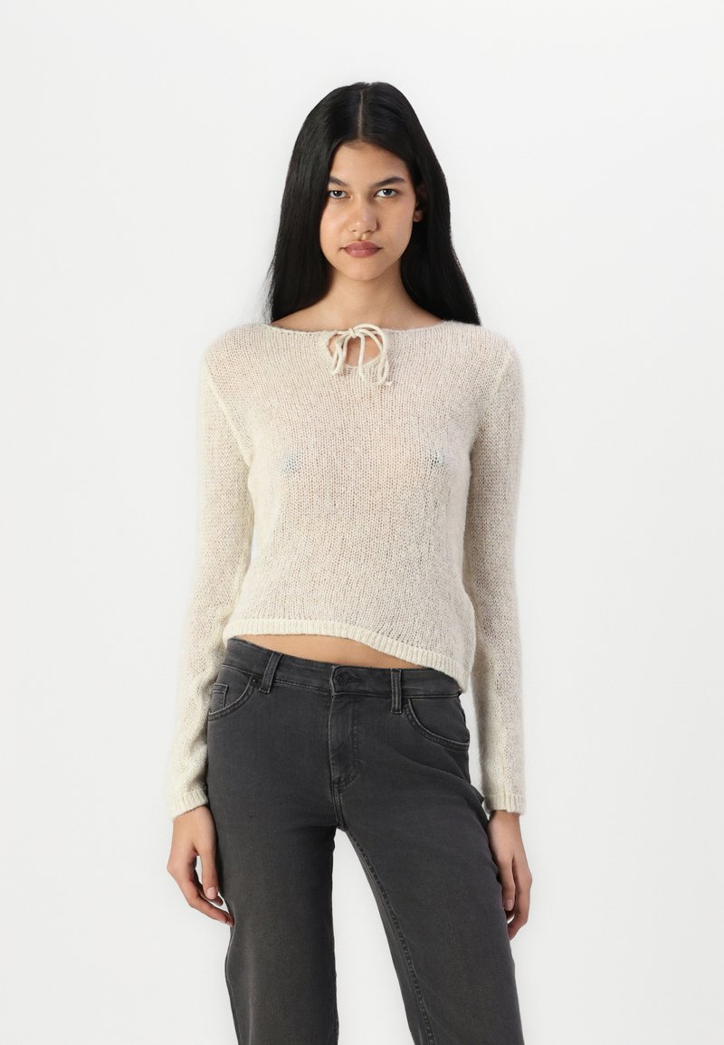 Lys beige strikket sweater med lange ærmer og rund halsudskæring, der har en sløjfedetalje foran; parret med mørkegrå jeans.