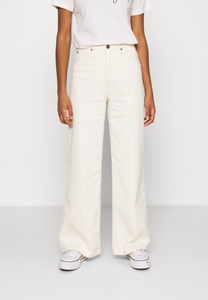 Osoba oblečená v bílé, široké kalhoty s vysokým pasem a předními kapsami a bílými teniskami, stojící před jednobarevným světlým pozadím.