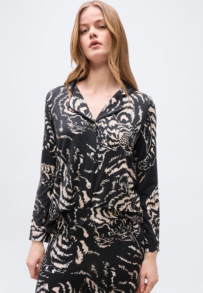 Νεαρή γυναίκα που φοράει μαύρο πουκάμισο με κουμπιά και παντελόνι με μπεζ και λευκό σχέδιο τίγρης σε απλό ανοιχτό φόντο.