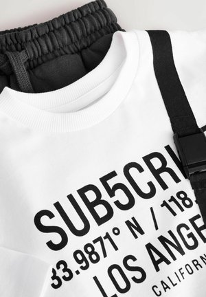 Hvid sweatshirt med sort tekst, der læser "SUB5CRV" og koordinater, udstyret med ribbestrikket krave og et bånd med spænde. Fastgjort er mørkegrå joggerstof.