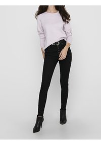 Pull léger lavande, jean skinny noir taille haute avec boutonnière devant, porté avec des bottines noires à talons. Fond uni.