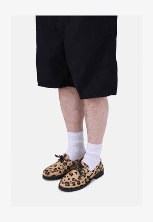 Zapatos con estampado de leopardo y suela negra, combinados con calcetines blancos y pantalones cortos negros. Los zapatos tienen cordones y un diseño de punta redonda.