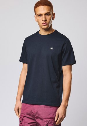 Joven con cabello corto, rizado y rojo, lleva una camiseta negra lisa y pantalones cortos cargo fucsia, de pie contra un fondo liso.