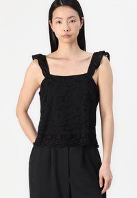 Black lace top with square neckline and ruffled straps, featuring floral patterns and textured accents at the hem. Paired with dark trousers.