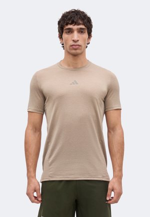 Homme portant un t-shirt de sport beige à manches courtes avec un petit logo sur la poitrine et un short vert foncé, debout devant un fond clair uni.