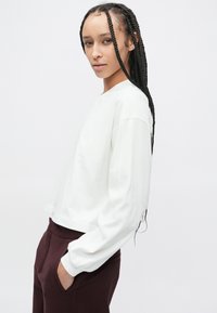 Biała bawełniana koszula z długim rękawem o luźnym kroju, okrągłym dekoltem i opuszczonymi ramionami, zestawiona z ciemnymi bordowymi spodniami.