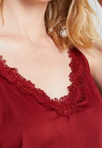 Burgundy cami med delikat blondekant langs halsudskæringen. Blødt, letvægtsstof med en glat tekstur og justerbare stropper.