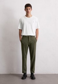 T-shirt blanc à manches courtes avec un petit logo, associé à un pantalon tapered vert olive. L'homme porte des chaussures noires à lacets, les mains dans les poches.