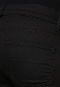 Svarta jeans med en slät textur och synlig sömnad. Har en bakficka med en lätt kurva och rena linjer.