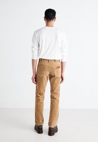 Hnědé kordové kalhoty se rovnými nohavicemi, detaily na zadních kapsách a hnědou koženou visačkou. V kombinaci s bílou vloženou košilí s dlouhým rukávem a tmavými botami.