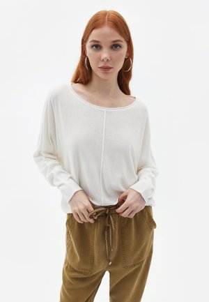 Weißes leichtes Langarmoberteil mit weitem Halsausschnitt und gerippten Bündchen, kombiniert mit braunen Cordhosen mit seitlichen Taschen und einem Kordelzug.