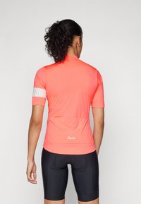 Kortermet sykkelskjorte i lys korallfarge, med hvite detaljer på ermene, pustende stoff og tre baklommer. Sorte sykkelshorts.