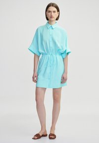 Une robe chemise à manches courtes en turquoise clair, avec une fermeture à bouton sur le devant, une taille ceinturée et un tissu doux avec une coupe décontractée.