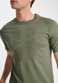 Oliwkowa koszula z krótkim rękawem wykonana z gładkiego materiału. Na przodzie znajduje się teksturowany wzór, a dekolt jest okrągły.