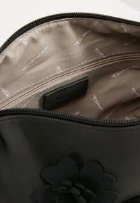 Schwarze Kosmetiktasche mit glatter Oberfläche und strukturiertem Blumenakzent. Das Innere ist mit einem beigen Futter versehen, das ein wiederholtes Logo-Muster aufweist, und bietet eine Reißverschlusstasche.