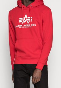 Sweat à capuche rouge avec un logo blanc portant le texte "ALPHA INDUSTRIES", poche kangourou et capuche avec cordon de serrage. Le matériau semble doux et durable.