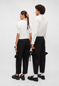 Duas modelos estão de costas uma para a outra, vestindo uma camisa branca de manga curta e calças largas pretas com laços laterais, combinadas com sapatos pretos robustos.