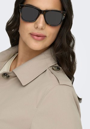 Nainen aaltoilevine tummine hiuksineen, jolla on ylisuurit mustat aurinkolasit ja beige trenssitakki, jossa on napitettu olkasauma, vaaleanharmaalla taustalla.