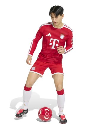 Jonge mannelijke voetballer in rode Bayern München-kit controleert een rode voetbal op een witte achtergrond.