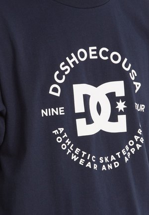 Námornícky modré bavlnené tričko s veľkým bielym grafickým logom a textom "DCSHOECOUSA" a "ATLETICKÁ SKATEBOARDOVÁ OBUV A ODEVY."
