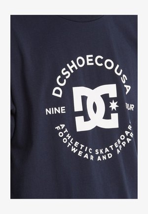 Námořnické modré bavlněné tričko se velkým bílým grafickým logem s textem "DCSHOECOUSA" a "ATLETICKÁ DESKOVÁ OBUV A OBLEČENÍ."