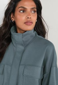 Soft, teal fabric top with a high collar, featuring two horizontal patch pockets and seam details for texture and design.