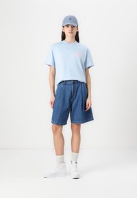 Ljusblå kortärmad t-shirt med rosa grafik, kombinerad med lösa denimshorts och vita hög-top sneakers, toppat med en blå basebollkeps.