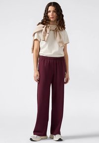 Lichtbeige T-shirt met een losse pasvorm, gecombineerd met bordeauxrode wijde broekspijpen. Een beige trui nonchalant om de schouders geknoopt. Sneakers maken de outfit af.