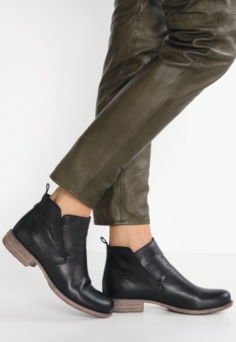 Bottes en cuir noir montant à la cheville avec des semelles marron portées avec un pantalon en cuir vert olive sur un fond blanc.