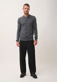 Šedý pletený cardigan s kulatým výstřihem, zapínáním na knoflíky, žebrovaným lemem, nošený přes černé kalhoty a černé boty. Jednoduchý, strukturovaný design.