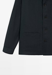 Veste noire à manches longues avec fermeture boutonnée et deux poches plaquées à l’avant, manches bordées de poignets, ourlet droit.