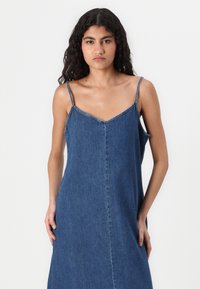 Dongerikjole med tynne stropper, V-hals og rett silhuett. Laget av blå denim, har et enkelt design uten synlige mønstre.