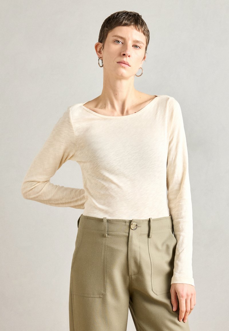 Ilgi rankoviai, šviesiai smėlinės spalvos marškiniai, pagaminti iš minkštos medžiagos su plačia apykakle, derinami su alyvuogių žalumo aukšto liemens kelnėmis, turinčiomis priekines kišenes.