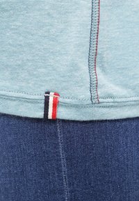 Ljusblå bomullstyg med röd söm och en etikett med vertikala ränder i röd, vit och marinblå nära fållen, i kombination med mörk denim.