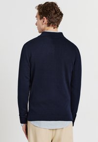 Mann von hinten gesehen, der einen dunkelblauen Langarmpullover über einem hellblauen Hemd und beigen Hosen vor einem schlichten Hintergrund trägt.