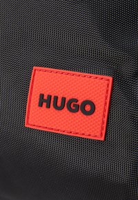 Juodas audinio maišas su tekstūruotu paviršiumi, kuriame yra išsiskirianti raudona stačiakampio formos detalė su iškilmingu juodu tekstu "HUGO."