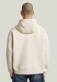 Man met een geschoren hoofd, gekleed in een beige hoodie en blauwe jeans, staand met zijn rug naar de camera tegen een eenvoudige achtergrond.