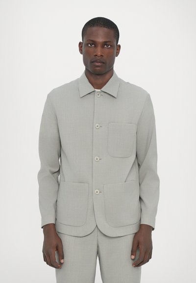 Ανοιχτό γκρι, υφασμάτινο σακάκι με μακριά μανίκια, γιακά, μία τσέπη και δύο μπροστινές πλαισιωτές τσέπες. Λείος υφασμάτινος φινίρισμα.