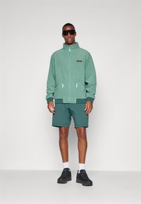Grønn fleecejakke med glidelåslommer, sammen med matchende teal shorts. Sorte sportssko og solbriller fullfører antrekket.