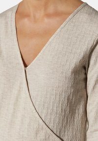 Beżowa tkanina teksturowana z dekoltem w kształcie litery V, z falistym wzorem i małym guzikiem przy dekolcie. Rękawy o dopasowanym kroju.