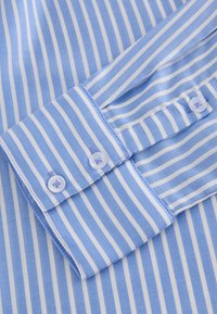 Mėlynos ir baltos juostelėmis margintos marškinių rankogalis su dviem baltais mygtukais. Audinys lygus, su smulkia tekstūra ir kontrastine siūlių detalė.