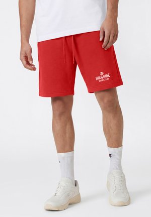 Uomo che indossa pantaloncini rossi con coulisse con la scritta "Ringside Boxing Club", calze bianche alla caviglia, scarpe da ginnastica bianche e una maglietta bianca corta sopra il ginocchio.