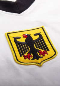 Black eagle with red beak and claws on a yellow shield patch sewn onto a white fabric with a black collar edge visible.